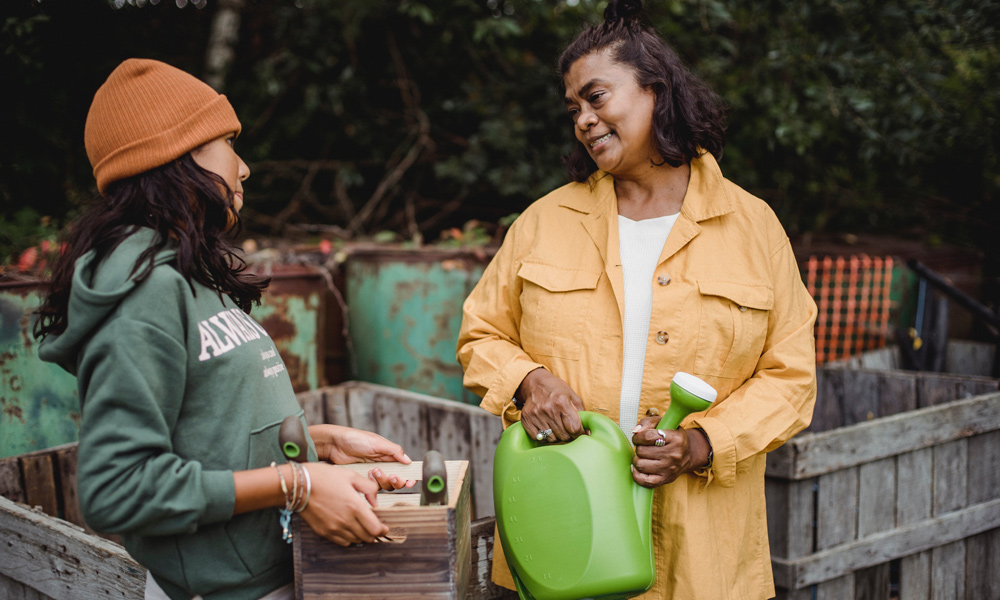 An woman of color holds a green watering can near a garden